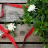 A wooden table holds red scissors, a spool of red ribbon, white flowers, a star-shaped tag with "Joy," and a green plant, evoking the warmth and spirit of Christmas trees.