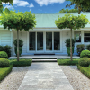 A single-story white house with a green roof and symmetrical landscaping featuring trimmed hedges, topiary, potted plants, and a stone path leading to the entrance showcases expert garden design.