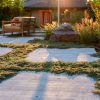 A backyard features a seating area with wooden chairs and a fire pit, surrounded by lush lawn alternatives and stone pavers, under the sunlight.